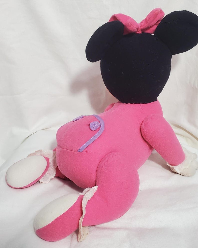 K&ouml;nnte beinhalten: Eine Pl&uuml;schpuppe von Minnie Mouse, die auf der Seite liegt. Die Puppe hat einen schwarzen Kopf mit gro&szlig;en Ohren und einer rosa Schleife. Sie tr&auml;gt ein rosa Outfit mit lila Akzenten und wei&szlig;er Verzierung an den F&uuml;&szlig;en.