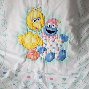May include: White fabric with a pattern of hearts and Big Bird and Cookie Monster appliqu&eacute;s. The characters are yellow and blue, with pink and orange accents. The fabric has a light blue and green diamond pattern with hearts.