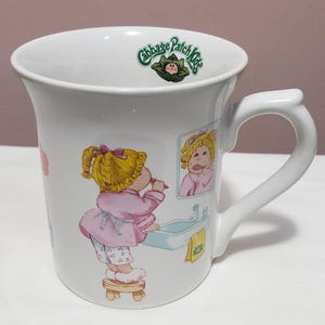 May include: White ceramic mug featuring a Cabbage Patch Kids design. The mug has a curved handle and a colorful illustration of a blonde girl brushing her teeth. The Cabbage Patch Kids logo is at the top.
