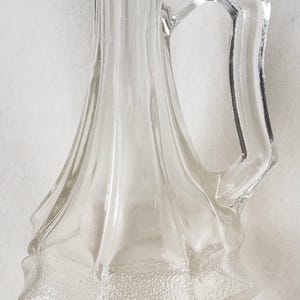 Peut inclure: Pichet en verre transparent avec une poignée. Le pichet a une base texturée en forme d'éclat d'étoile et un corps élancé et cannelé. La poignée est angulaire et géométrique. Le pichet est probablement destiné à servir des boissons.