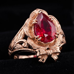 May include: A rose gold ring featuring a large, pear-shaped, faceted red gemstone. The ring's setting is a detailed floral design with a bow accent. The band has an intricate leaf pattern, creating an elegant and romantic aesthetic.