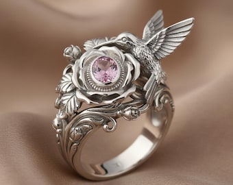 Könnte beinhalten: Silberner Ring mit einem Kolibri mit ausgebreiteten Flügeln über einer Rose mit einem rosa Edelstein. Der Ring hat detaillierte florale Muster und eine polierte Oberfläche. Das Design ist von der Natur inspiriert.