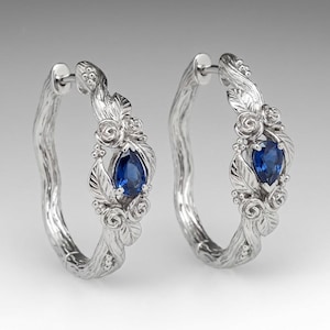 May include: A pair of silver hoop earrings featuring a textured branch-like design adorned with sculpted roses and leaves. Each earring is centered with a single pear-shaped blue sapphire gemstone held in a prong setting.