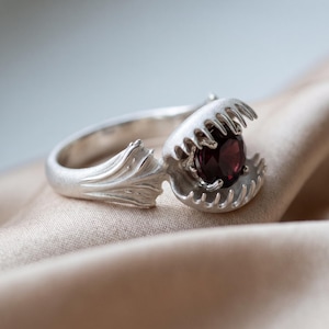 Op de afbeelding: Een zilveren ring met een zetting in de vorm van een Venusvliegenvanger met gekartelde randen. Het midden van de plantachtige structuur bevat een ronde, dieprode edelsteen. De ringband heeft een gestructureerd, organisch ontwerp als bladeren.