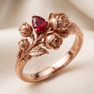 Può includere: Un anello in oro rosa con un design floreale. Presenta tre rose e una gemma rosso scuro a forma di pera. La fascia dell'anello è liscia e le rose hanno petali e foglie dettagliati.