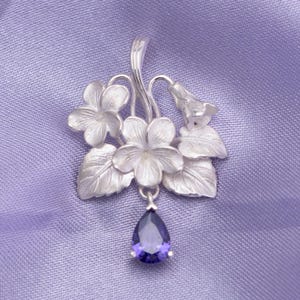 Puede incluir: Un colgante de plata con un grupo de flores y hojas esculpidas con acabado mate. Una única piedra preciosa morada, facetada y en forma de pera, cuelga de la parte inferior del arreglo floral, que está sujeto a una anilla de plata en la parte superior.