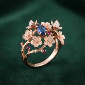 May include: Rose gold ring featuring a central oval blue gemstone, surrounded by delicate floral and butterfly designs. Small red gemstones accent the branches of the ring. The ring's intricate details and warm tones create an elegant and nature-inspired aesthetic.