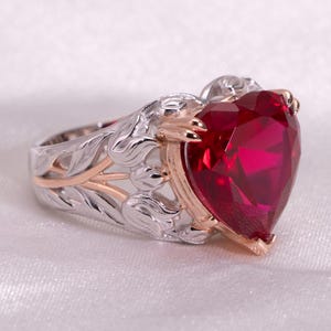 Pode incluir: Um anel com uma grande pedra preciosa vermelho-escura em forma de coração, montada numa banda de metal bicolor. A banda é feita de metal prateado com padrões intrincados de folhas e vinhas, acentuada com detalhes em metal ouro rosa.