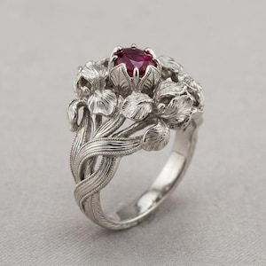 May include: A silver ring featuring a floral design with a central, round, deep red gemstone. The ring's band is intricately detailed with interwoven lines, and the setting is adorned with sculpted flower petals.