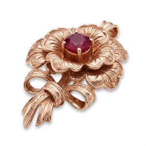 Pode incluir: Pingente em ouro rosa com design floral e uma gema central redonda de cor vermelho escuro. O pingente inclui um laço detalhado e elementos florais, mostrando um artesanato intrincado. Projetado para ser usado como colar.