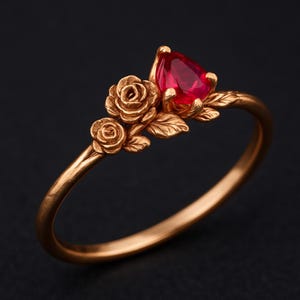 May include: A gold-colored ring featuring a pear-shaped, vibrant red gemstone. The ring is adorned with delicate rose and leaf details, creating a floral design. The ring's band is smooth and polished, set against a dark background.
