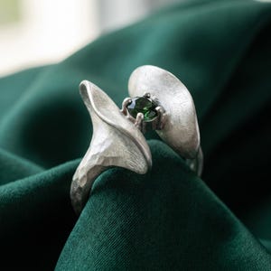 Op de afbeelding: Een zilveren ring met een getextureerde, gehamerde metalen band die zich splitst in twee gebogen, bloembladachtige vormen. Een enkele, ronde, donkergroene edelsteen is in het midden tussen de twee zilveren elementen gezet, wat een bloemachtig uiterlijk creëert.