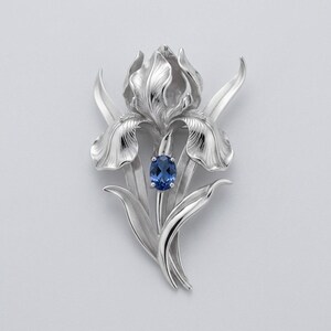 Pode incluir: Broche prateado em forma de flor de íris. O broche apresenta detalhes de pétalas e folhas, com uma pedra preciosa azul oval no centro. O design é elegante e botânico, adequado para ocasiões formais ou como acessório decorativo.