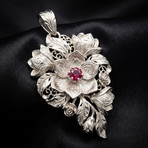 May include: A silver pendant featuring a floral design with a central ruby gemstone. The pendant showcases detailed leaf and flower carvings, with a bail for a chain. The jewelry is set against a black background.