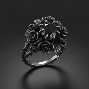 Op de afbeelding: Een ring van donkergrijs metaal met een centrale, rondgeslepen zwarte edelsteen in een klauwzetting. De ringband en zetting zijn vormgegeven als een gedetailleerd arrangement van driedimensionale rozen, bladeren en doornige ranken.