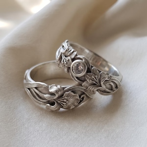Könnte beinhalten: Zwei Silberringe mit detaillierten floralen Reliefschnitzereien. Ein Ring ist mit einem einzelnen runden, klaren, facettierten Edelstein besetzt. Das Metall hat ein mattes, gebürstetes Finish mit oxidierten Vertiefungen, die die Blütenstrukturen betonen.