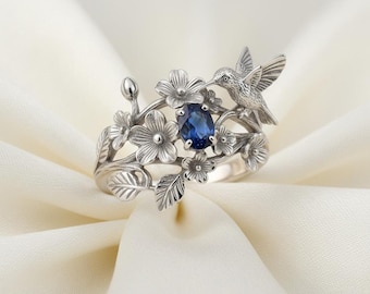May include: A silver ring featuring a hummingbird and floral design. The ring has a central, oval-shaped, deep blue gemstone. The hummingbird is perched on a branch with detailed flowers and leaves, creating a nature-inspired aesthetic.