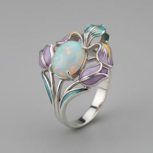 Puede incluir: Anillo de plata con una piedra de ópalo ovalada, rodeada de detalles florales esmaltados. Las flores están pintadas en morado, amarillo y turquesa. El diseño del anillo está inspirado en los iris.