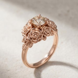 Pode incluir: Anel de ouro rosa com um grande diamante de corte quadrado. A banda do anel é adornada com rosas e folhas esculpidas, criando um design floral. O anel é exibido sobre um fundo neutro, destacando seus detalhes intrincados.
