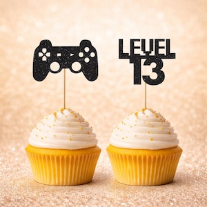 Może przedstawiać: Dwie babeczki z białym lukrem i złotymi posypkami. Jedna babeczka ma czarną nakładkę kontrolera, druga czarną nakładkę "LEVEL 13". Babeczki stoją na złotym, błyszczącym tle.