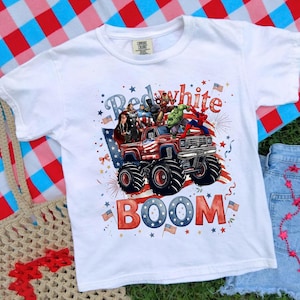 Op de afbeelding: Wit T-shirt met een rood, wit en blauw monster truck ontwerp met superhelden. De tekst op het shirt luidt "Red White Boom" met ster- en vlagaccenten, perfect voor patriottische evenementen.