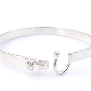 Op de afbeelding: Een zilverkleurige metalen armband met een glad, plat ontwerp. Het ene uiteinde van de armband eindigt in een kleine, afgeronde haak, terwijl het andere uiteinde een lusdetail heeft met drie horizontale ribbels voor een veilige sluiting.