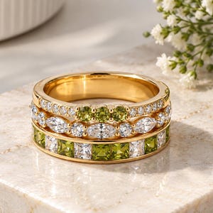 May include: A stack of three gold rings. The top ring features small round green stones. The middle ring has alternating marquise and round clear stones. The bottom ring displays a channel setting with alternating square green and clear stones.