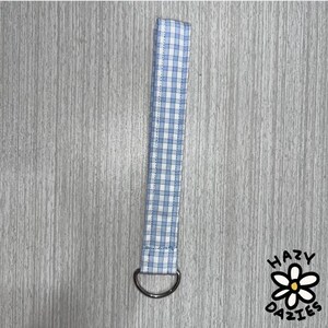 Puede incluir: Un llavero de muñeca con estampado de cuadros vichy azul y blanco con una anilla en D plateada. El llavero es de tela y está diseñado para llevarlo en la muñeca. Un accesorio práctico y elegante.