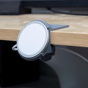 May include: A gray plastic mount holds a circular white wireless charging pad at the edge of a flat surface. The mount features a flat extension that rests on top of the surface, while the charging pad is positioned vertically on the front.