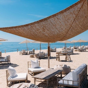 Può includere: Una scena sulla spiaggia con mobili in bambù, cuscini bianchi e un grande parasole intrecciato. La scena comprende sedie a sdraio, tavoli e ombrelloni su una spiaggia sabbiosa. L'oceano e il cielo blu sono visibili sullo sfondo.