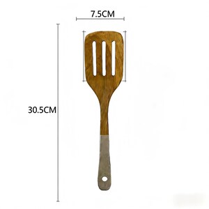 May include: A wooden spatula with a slotted head, measuring 7.5 cm wide and 30.5 cm long. The handle has a silver metal base with a hole for hanging. The spatula is made of light brown wood.