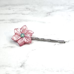 May include: A pink flower hair clip with a light blue gem in the center. The clip is made of metal and has a simple design.