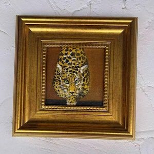May include: A framed painting of a jaguar's face, with a gold-colored frame. The jaguar has yellow fur with black spots and green eyes. The background is a dark brown color.