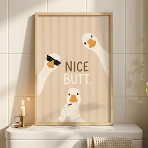 Op de afbeelding: Een ingelijste wandprint met drie witte ganzen op een beige gestreepte achtergrond. Eén gans draagt een zwarte zonnebril, een andere houdt een rol toiletpapier vast en de tekst NICE BUTT. staat in het bruin in het midden gedrukt.