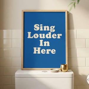 Könnte beinhalten: Ein rechteckiges Wandbild in einem hellen Holzrahmen mit einem leuchtend blauen Hintergrund und dem Text "Sing Louder In Here", der in einer fetten, cremefarbenen, abgerundeten Serifenschrift gedruckt ist. Das Kunstwerk ist vertikal ausgerichtet.