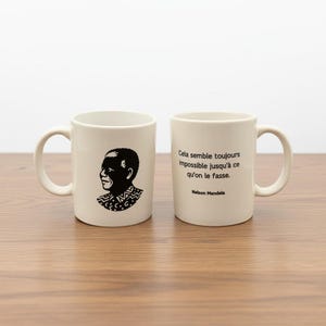 Könnte beinhalten: Zwei weiße Keramikbecher. Ein Becher zeigt eine schwarze Silhouette von Nelson Mandela und der andere ein Zitat von Nelson Mandela in schwarzem Text. Die Becher haben einen gebogenen Griff.