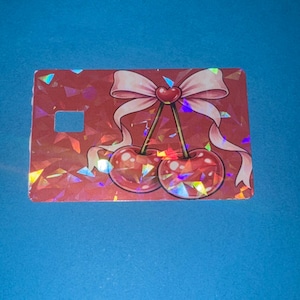 May include: A red, rectangular card with a holographic, geometric pattern. The card features a pink bow with a heart in the center, and two cherries. The background is a solid blue.