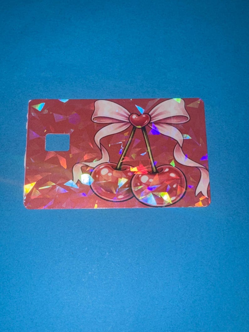 May include: A red, rectangular card with a holographic, geometric pattern. The card features a pink bow with a heart in the center, and two cherries. The background is a solid blue.