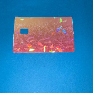 May include: A rectangular, iridescent card with a cut-out square on the left side. The card has a gradient of colors, transitioning from orange to pink, with scattered, colorful geometric shapes.