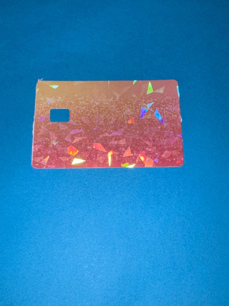 May include: A rectangular, iridescent card with a cut-out square on the left side. The card has a gradient of colors, transitioning from orange to pink, with scattered, colorful geometric shapes.