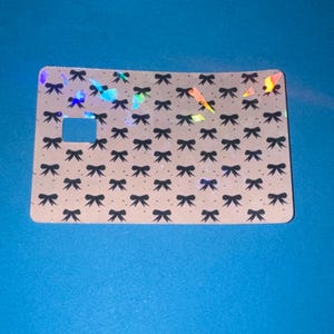 May include: A rectangular card with a light tan background, covered in a pattern of small black bows. The card has a square cutout and iridescent holographic accents. The card is set against a blue background.