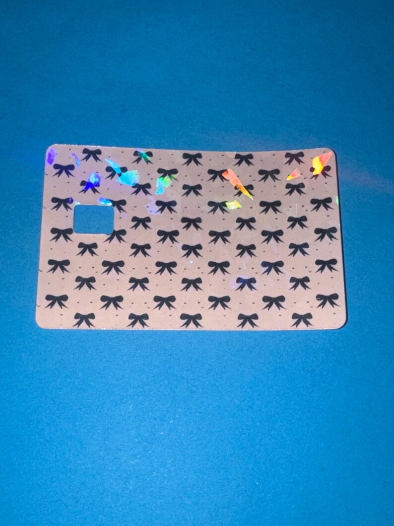 May include: A rectangular card with a light tan background, covered in a pattern of small black bows. The card has a square cutout and iridescent holographic accents. The card is set against a blue background.