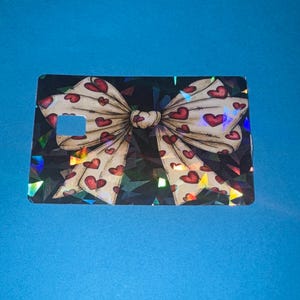 May include: A rectangular card with a bow design. The bow is cream-colored with red heart patterns. The background is black with a geometric pattern. The card has a small rectangular cutout on the left side.