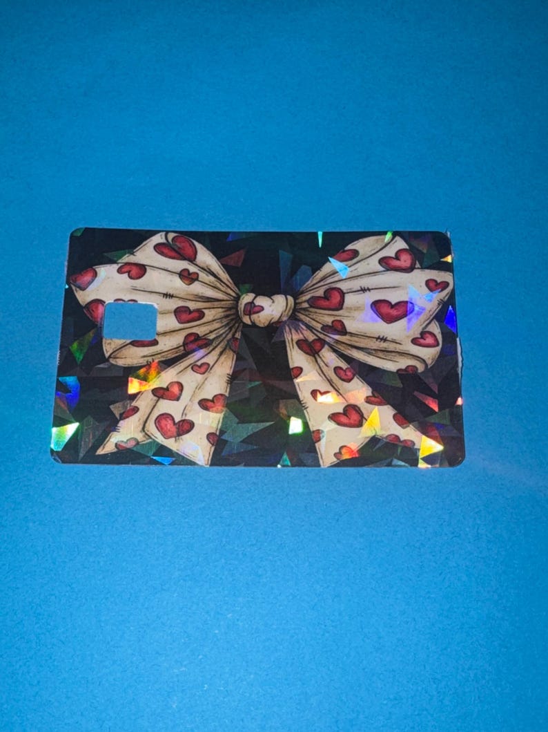 May include: A rectangular card with a bow design. The bow is cream-colored with red heart patterns. The background is black with a geometric pattern. The card has a small rectangular cutout on the left side.