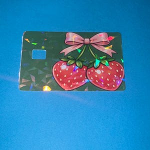 May include: A green card with a strawberry design. Two red strawberries with green stems and leaves are connected by a pink bow. The card has a holographic, geometric pattern and a rectangular cutout.