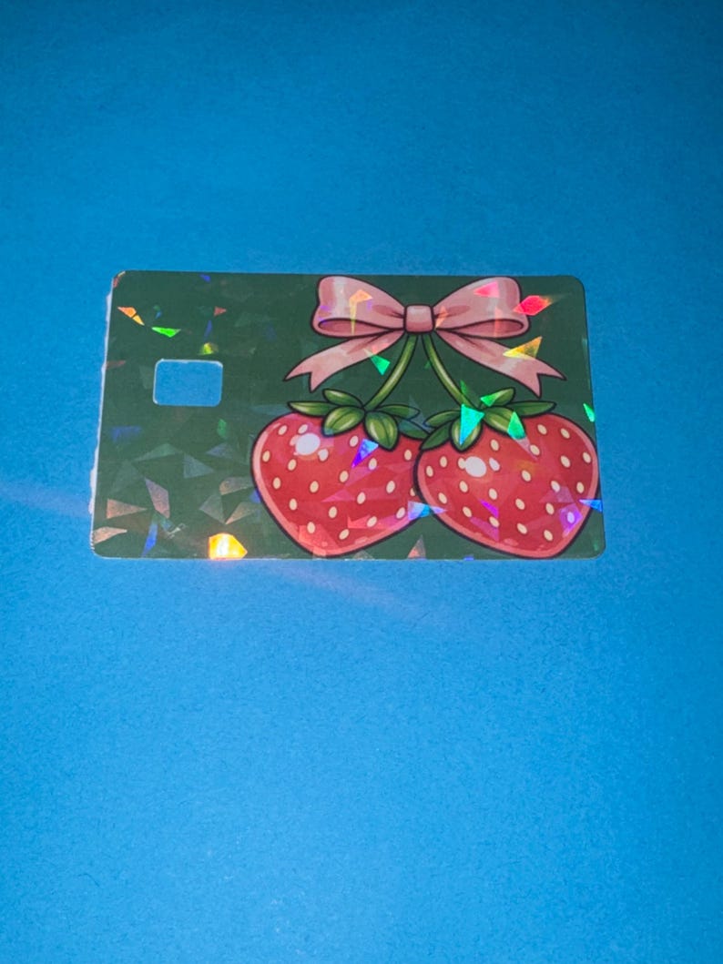 May include: A green card with a strawberry design. Two red strawberries with green stems and leaves are connected by a pink bow. The card has a holographic, geometric pattern and a rectangular cutout.