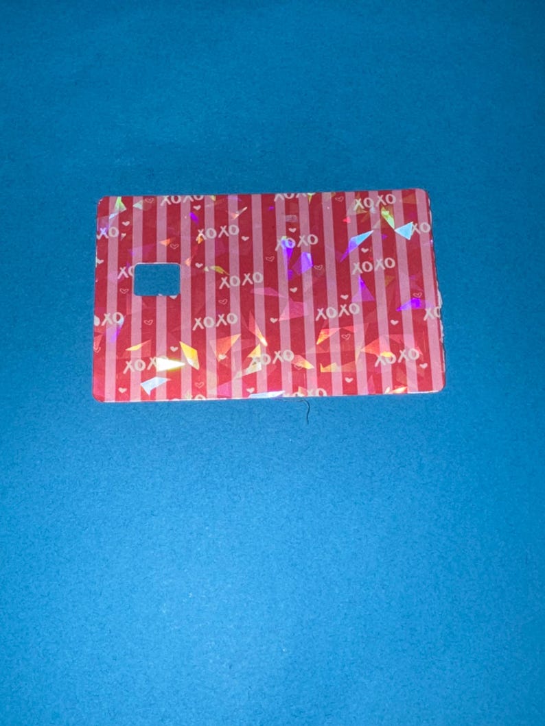 May include: A rectangular card with a red and white striped pattern, featuring the text "XOXO" and small heart shapes. The card has a rectangular cutout and holographic accents, set against a blue background.