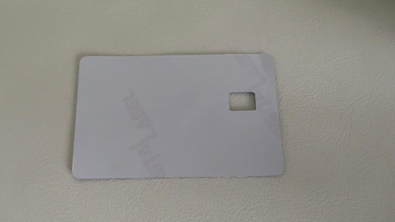 Puede incluir: Una tarjeta rectangular blanquecina con un recorte cuadrado cerca del borde derecho. La tarjeta tiene esquinas redondeadas y parece estar hecha de un material liso y mate. El fondo es una superficie texturizada de color claro.