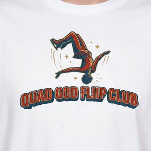 May include: White t-shirt featuring a graphic of a person doing a flip in red and orange. The text "QUAD GOD FLIIP CLUB" is printed in a retro font in blue and red. The shirt is made of a soft material.
