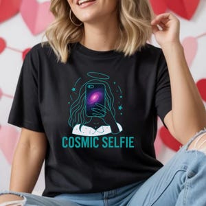 May include: Black t-shirt with a teal graphic of a person taking a selfie with a galaxy on their phone. The text "COSMIC SELFIE" is below the graphic. The design includes a halo and stars.